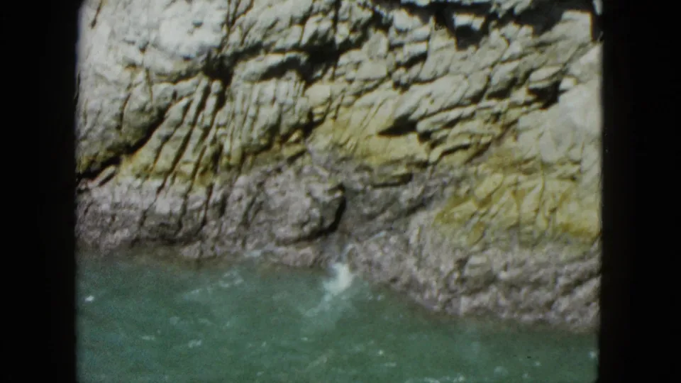 1962 Acapulco frame showing turquoise water and small surf against a rugged rock base beneath sunlit stone.