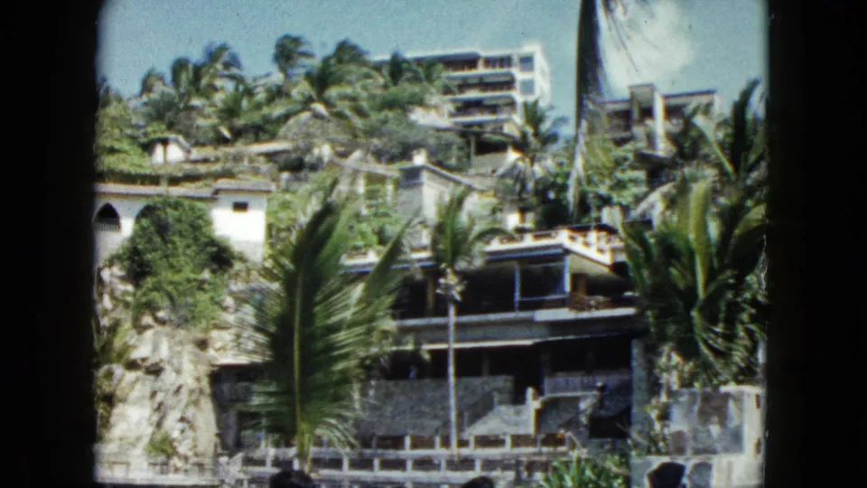 1962 Acapulco home-movie view of palm-lined hillside buildings, rocky terrain, and stone terraces.