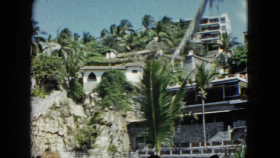 1962 Acapulco frame showing a rocky palm-covered hillside with buildings partly hidden by tropical foliage.
