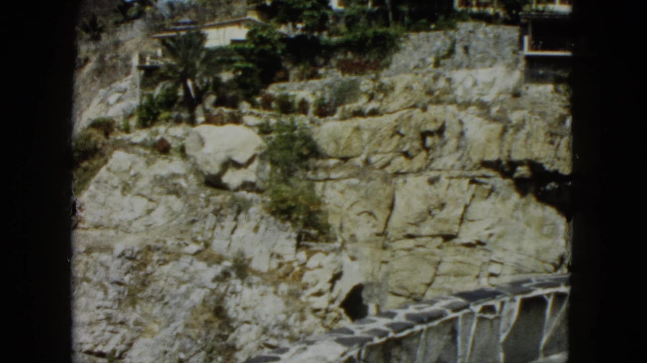 Close-up of a rocky limestone cliff face with white buildings and tropical palm trees visible along the upper ridge.