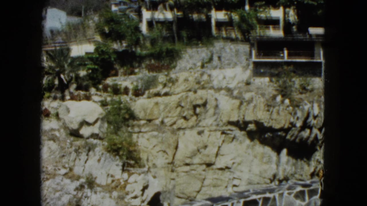 Rocky cliff face with white terraced buildings and dense palm vegetation at the summit, slight pan from previous frame.