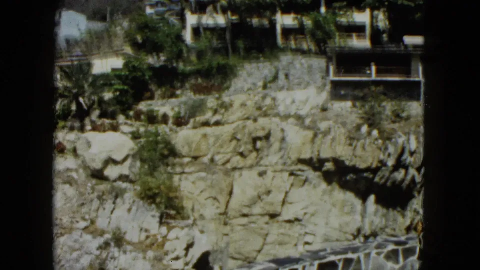 8mm frame of a cliff face in Acapulco, Mexico, circa 1962, showing white resort buildings and tropical palms at the crest with a railing structure visible.