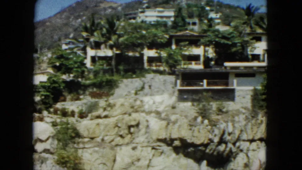8mm frame of cliffside modernist villas in Acapulco, Mexico, circa 1962, with lush tropical vegetation, rocky cliff base, and a small human figure at the lower right edge.