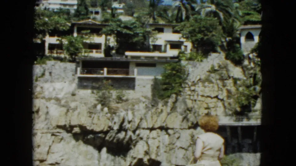8mm frame of cliffside resort architecture in Acapulco, Mexico, circa 1962, with tropical palms, rocky cliff base, and a small human figure visible at the frame edge.