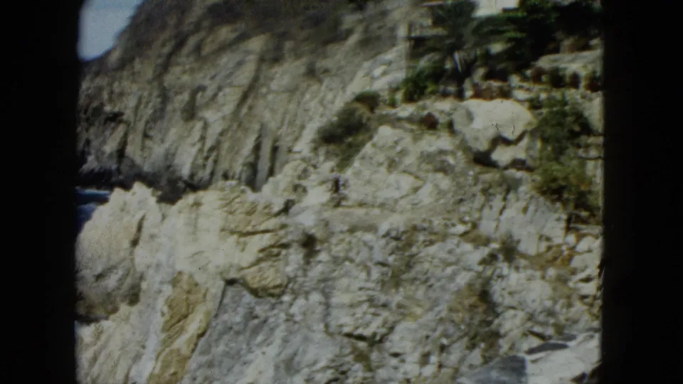 Rocky coastal cliff face with scrub growth in crevices, ocean at upper left, possible white structure at upper right, Acapulco circa 1962