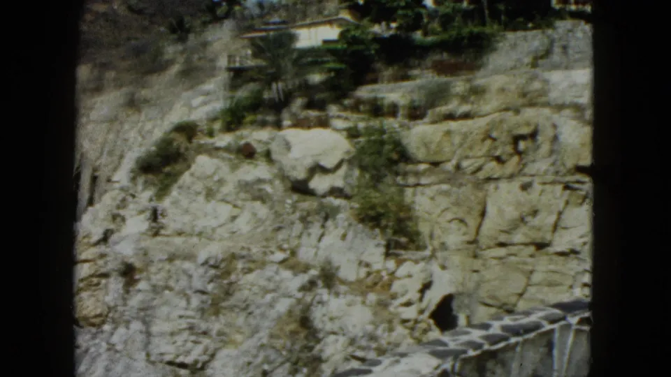 Acapulco coastal cliff face with scrub growth, glimpse of white building at upper right, and railing at lower right corner, circa 1962