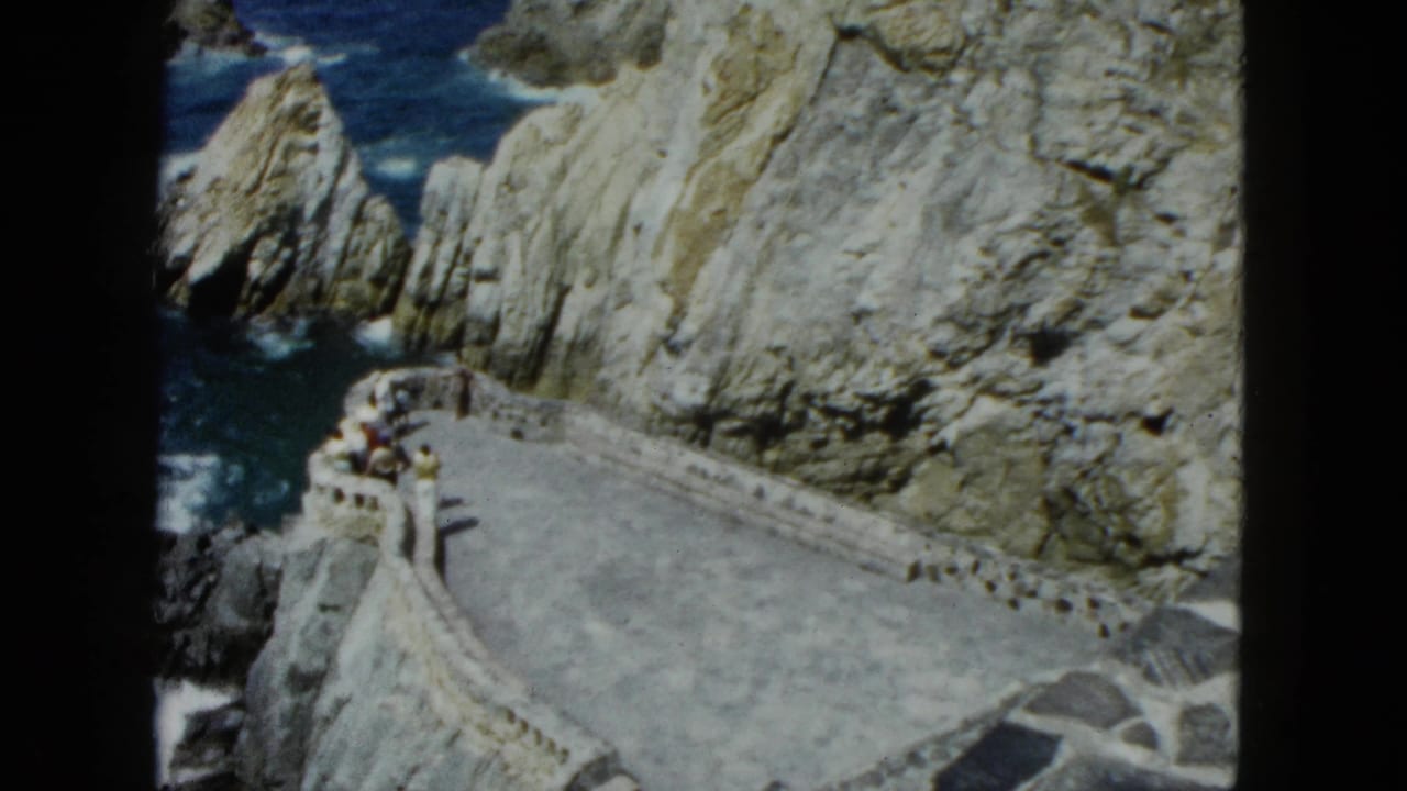 Elevated view of a curved stone walkway along a rugged cliff above blue ocean, with small figures of visitors visible.
