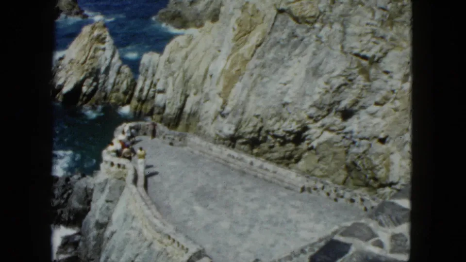 Elevated 8mm frame showing a curved stone promenade with low balustrade along a dramatic cliff, visitors visible, Acapulco area, circa 1962.