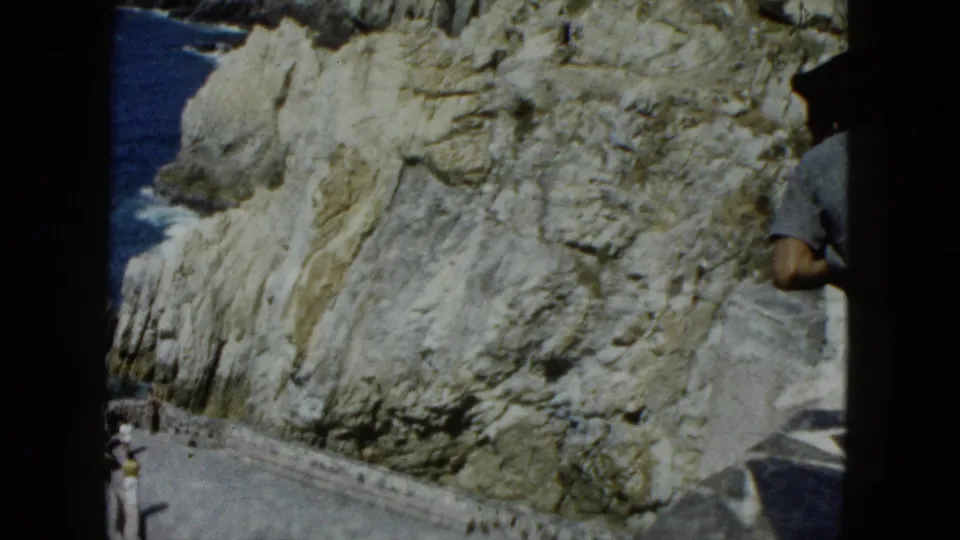 8mm frame: man in gray short-sleeve shirt at frame edge, dramatic stratified cliff rock and blue Pacific ocean in background, Acapulco area, circa 1962.