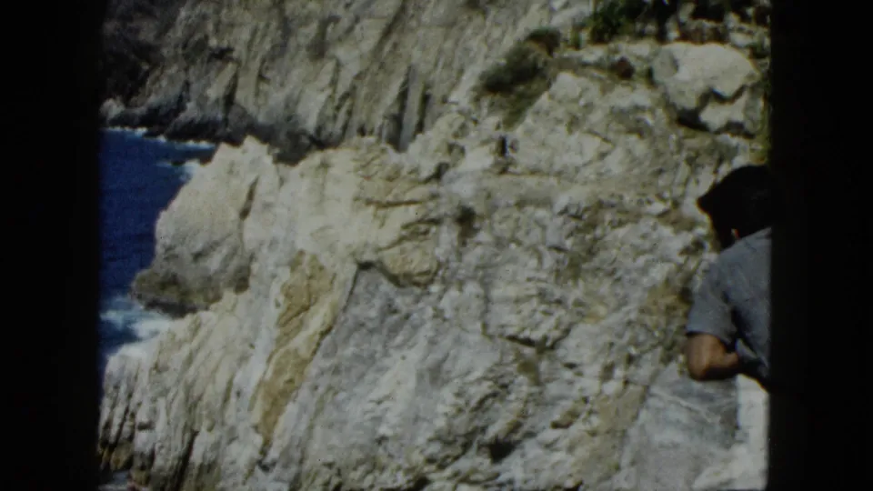 8mm frame with partial figure at frame edge, large rugged stratified cliff face, blue Pacific ocean to the left, Acapulco coastline, circa 1962.