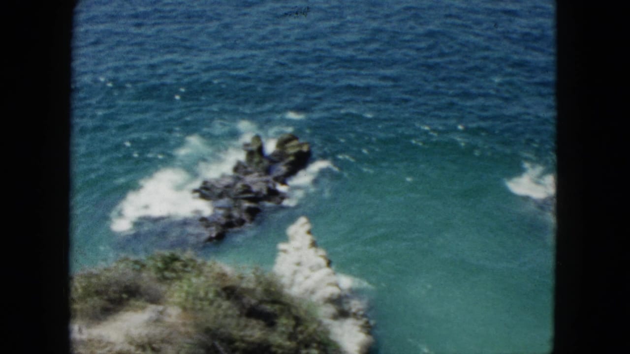 1962 Acapulco home-movie frame showing turquoise water, white surf, dark offshore rocks, and a pale cliff edge from an elevated viewpoint.