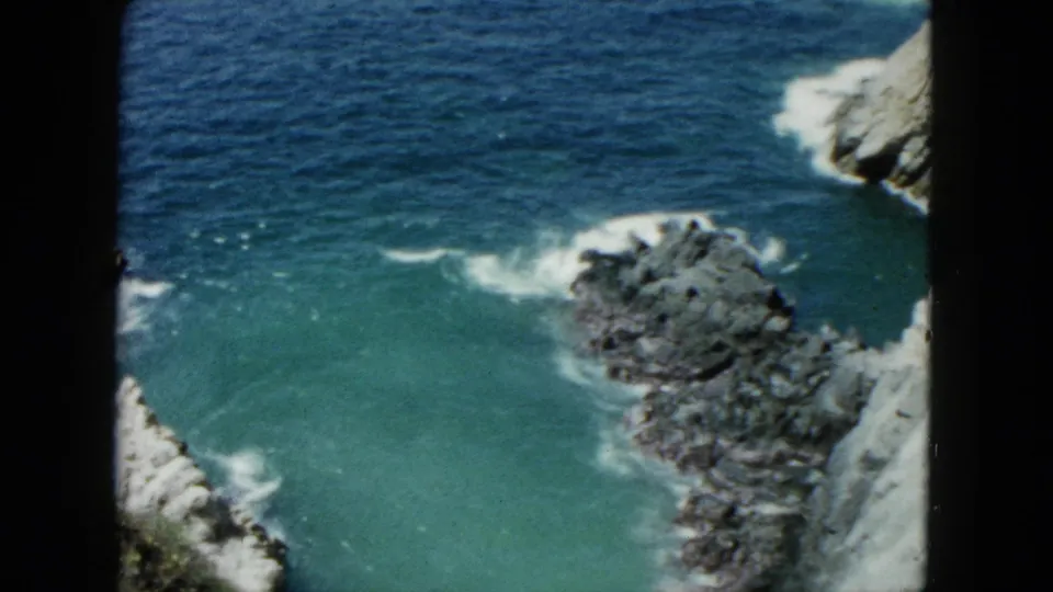 1962 Acapulco coastal view with blue-green water, white wave lines, jagged rocks, and pale cliff faces along the edge of the frame.