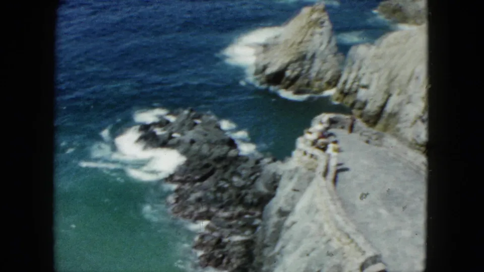 1962 Acapulco home-movie view of a stone cliffside walkway, low wall, pale rock face, turquoise water, and distant visitors near the overlook.