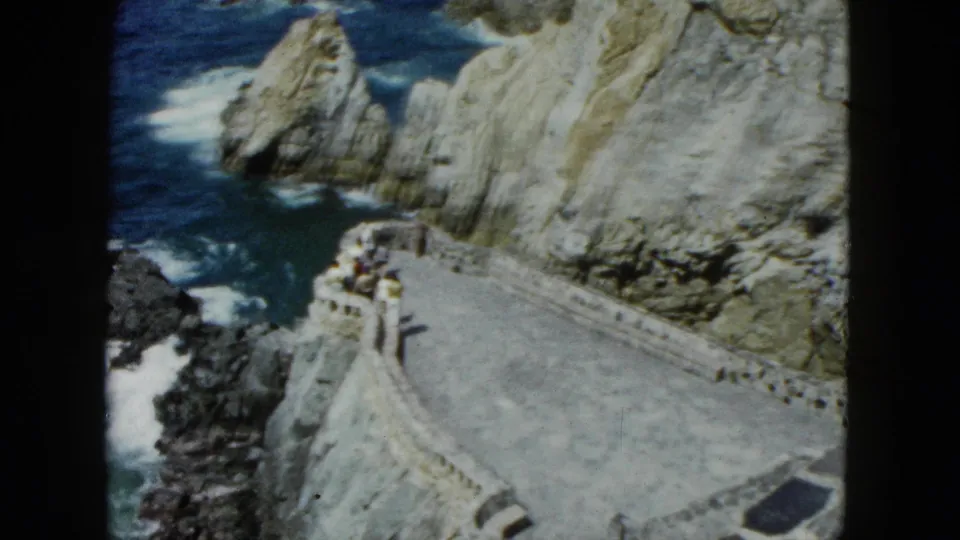 1962 Acapulco frame showing a stone overlook path beside pale coastal cliffs, dark rocks below, white surf, and small distant people along the wall.