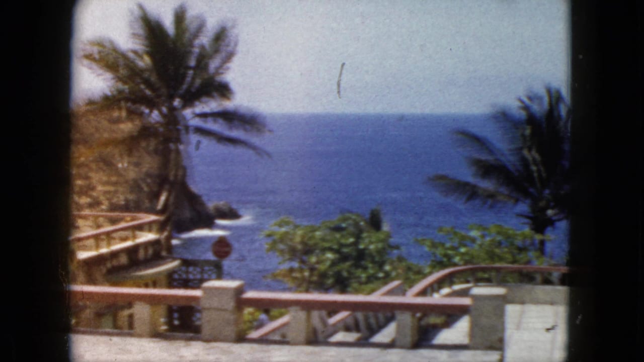 1962 Acapulco home-movie frame showing palm trees, a painted terrace railing, tropical foliage, and the Pacific horizon beyond a cliffside view.