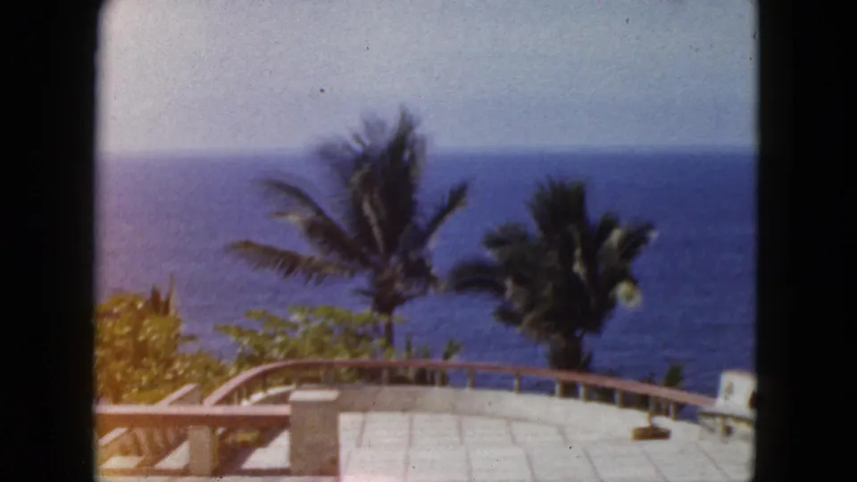 1962 Acapulco terrace shot with pale pavement, palms, dense coastal greenery, and the blue Pacific visible beyond the overlook.