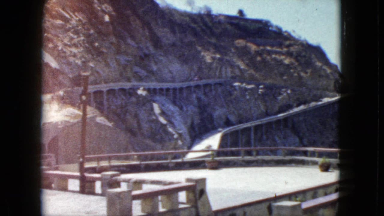 1960s 8mm home-movie frame showing a concrete observation terrace and cliffside road, likely Acapulco, Mexico, circa 1962; lens flare from bright sky.