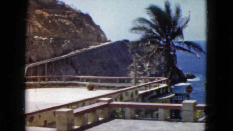 1960s 8mm frame after camera pan, revealing Pacific Ocean, palm tree, and stop sign alongside the terrace railing, Acapulco, Mexico, circa 1962.