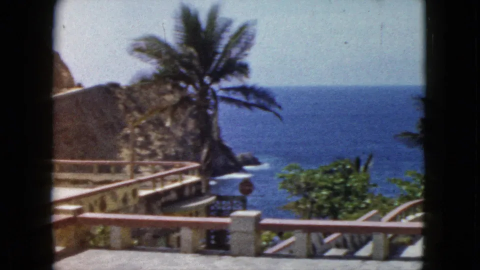 Final pan frame, 1960s 8mm, showing Acapulco Pacific coastline with palm tree, terrace railing, stop sign, and open ocean, circa 1962.
