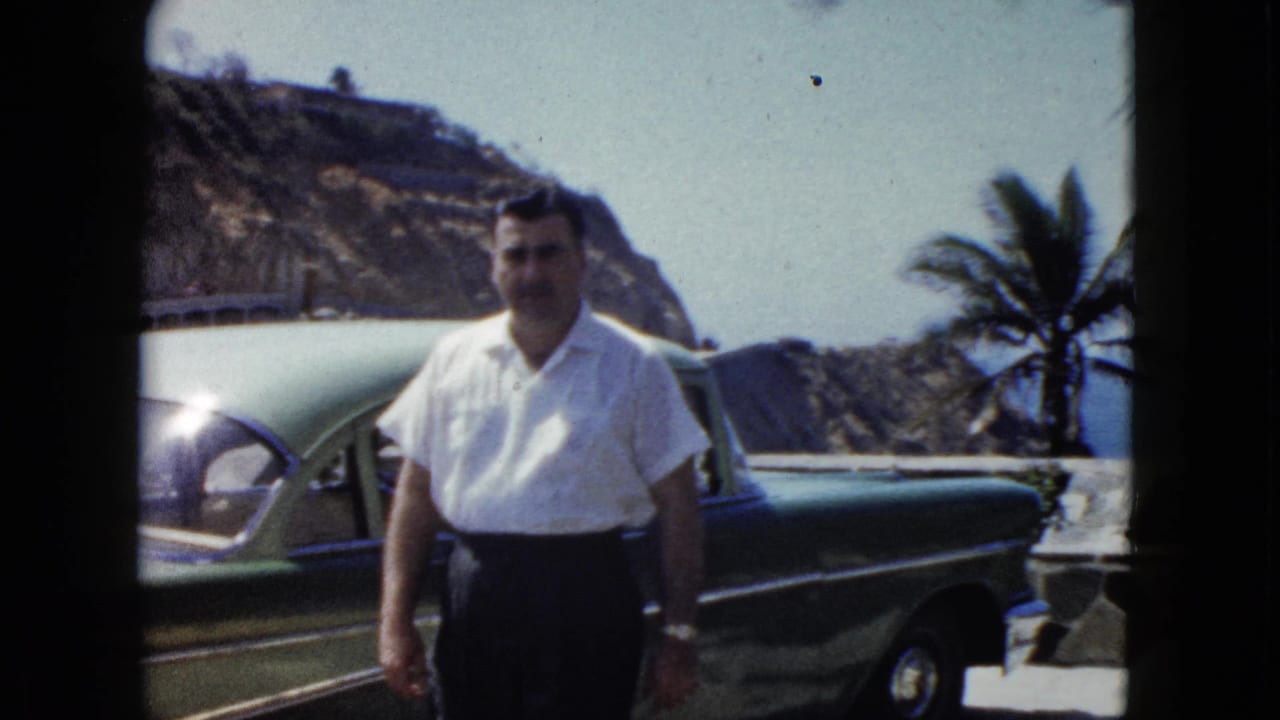 Man in white short-sleeved shirt and dark trousers poses beside a green classic American car at an Acapulco coastal overlook, circa 1962.