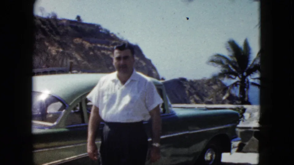 Man in white short-sleeved shirt and dark trousers poses beside a green classic American car at an Acapulco coastal overlook, circa 1962.