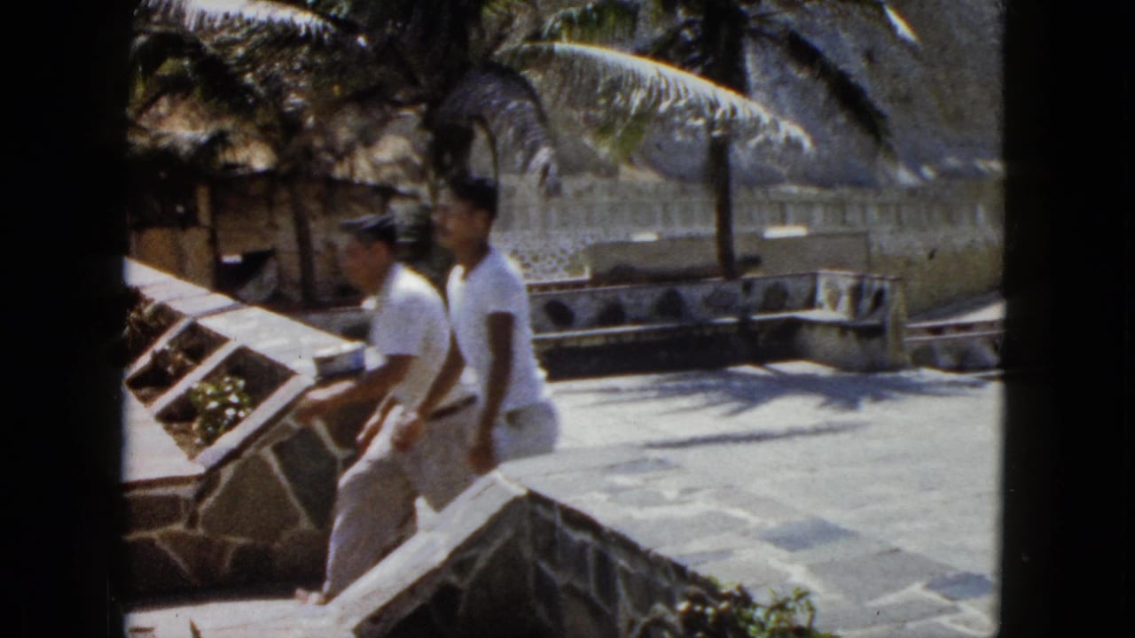 Two men in white shirts on a stone terrace at what appears to be an Acapulco coastal viewpoint, with a rocky hillside and stone wall in the background, circa 1962.