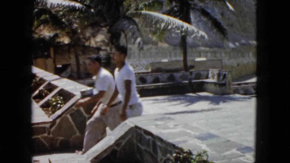 Two men in white shirts on a stone terrace at what appears to be an Acapulco coastal viewpoint, with a rocky hillside and stone wall in the background, circa 1962.