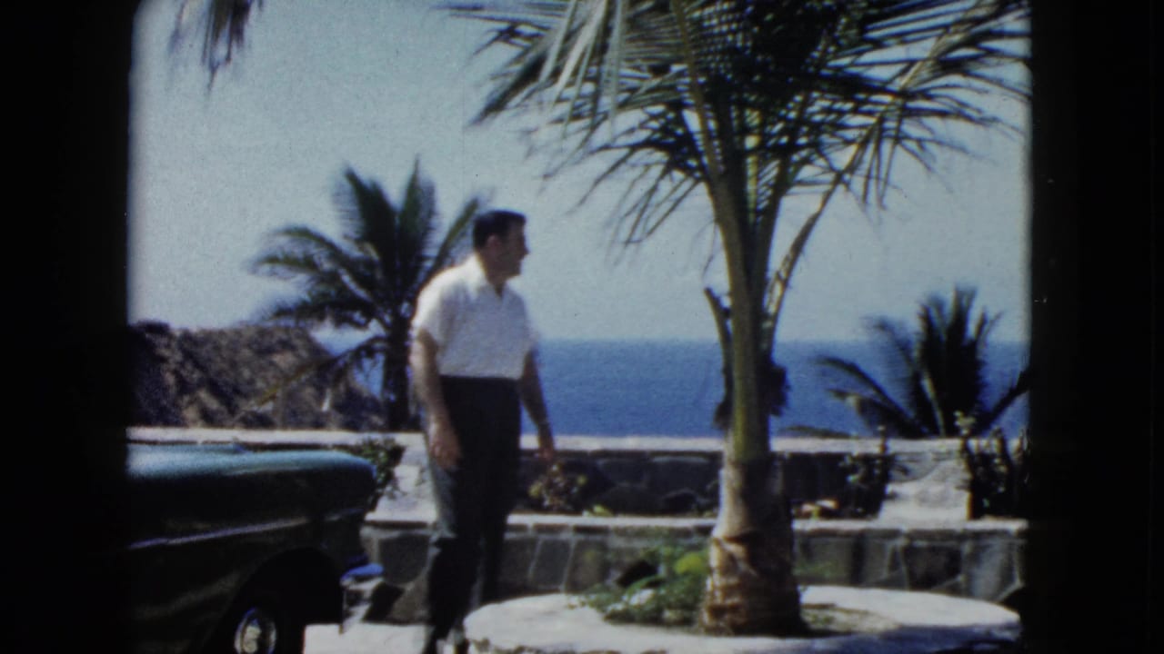 Man in white short-sleeved shirt stands sideways on a stone terrace with a palm tree and ocean behind him, dark vintage sedan to the left.