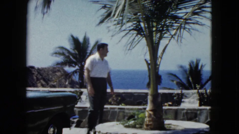 Middle-aged man in white shirt stands in profile on a stone oceanfront terrace in what appears to be Acapulco, Mexico, circa 1962, with a dark vintage automobile and palm trees visible.