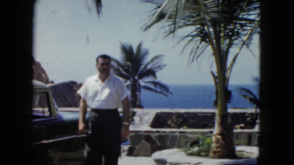 Middle-aged man with mustache in white shirt approaches camera on a stone terrace overlooking the Pacific, Acapulco area, circa 1962; dark vintage sedan visible at left.