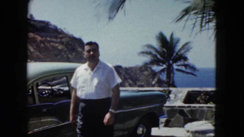Middle-aged man with mustache faces camera at close range on a stone terrace, Acapulco area, circa 1962; a dark late-1950s sedan, palm trees, and a rocky coastal headland frame the scene.