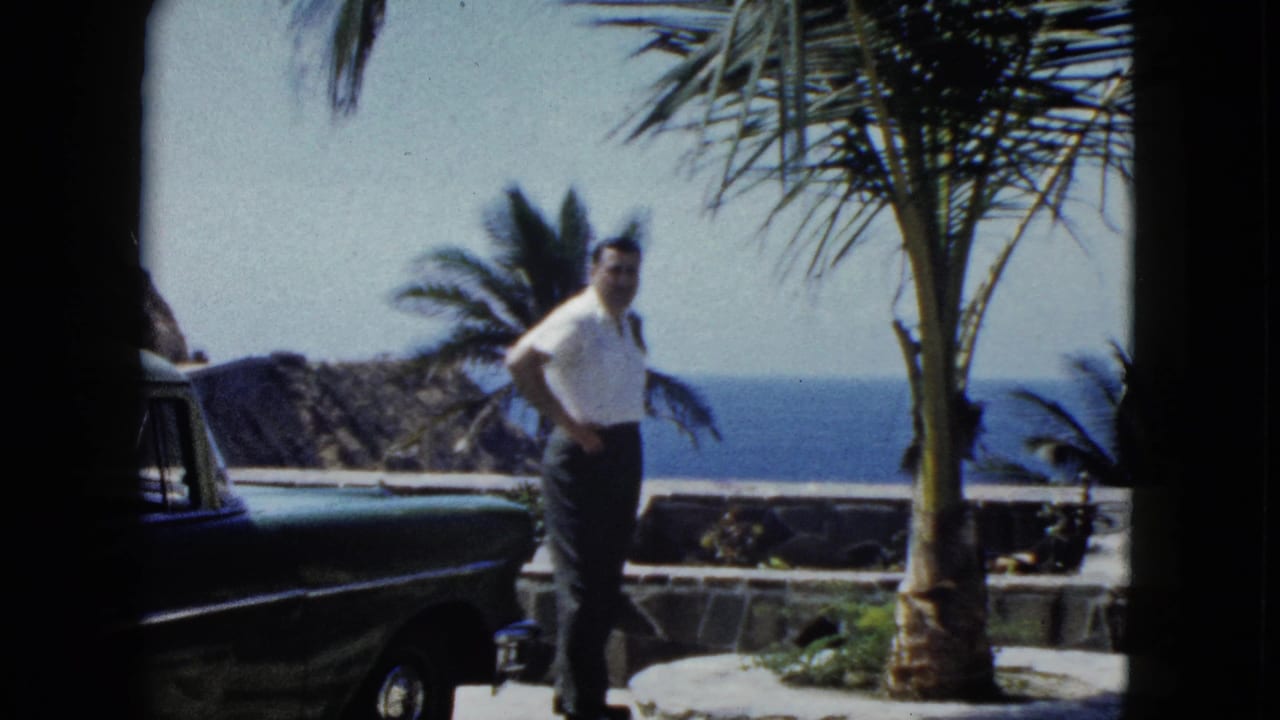 Man in white shirt stands beside a dark vintage American car with palm trees and ocean in the background.