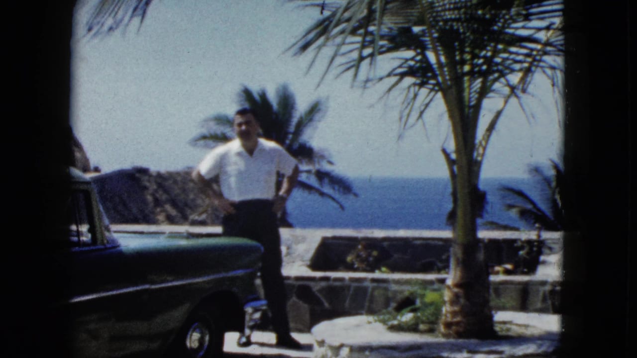 Man with hands on hips stands beside a large-finned vintage car; palm trees and blue ocean visible behind him.