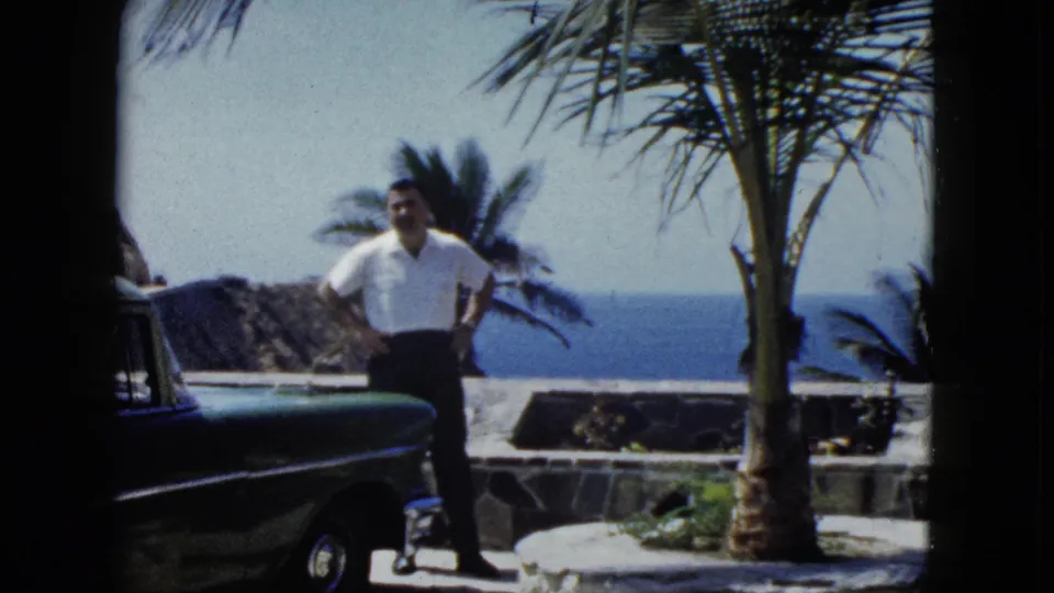 Man posing with hands on hips beside a late-1950s or early-1960s American automobile at a coastal terrace, Acapulco, circa 1962.