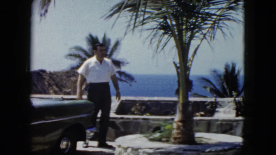Man stepping away from vehicle beneath a palm tree, stone terrace and ocean behind, Acapulco area, circa 1962.