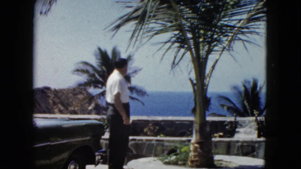 Man turning near a palm tree beside a vintage automobile, with rocky coastal cliffs and blue ocean behind, Acapulco, circa 1962.