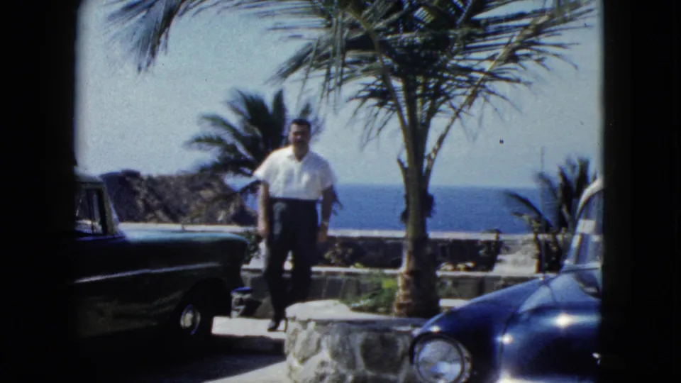 Tourist stands on a stone coastal terrace in Acapulco, Mexico, circa 1962, flanked by a large dark blue American-style automobile and a palm tree, ocean visible beyond.