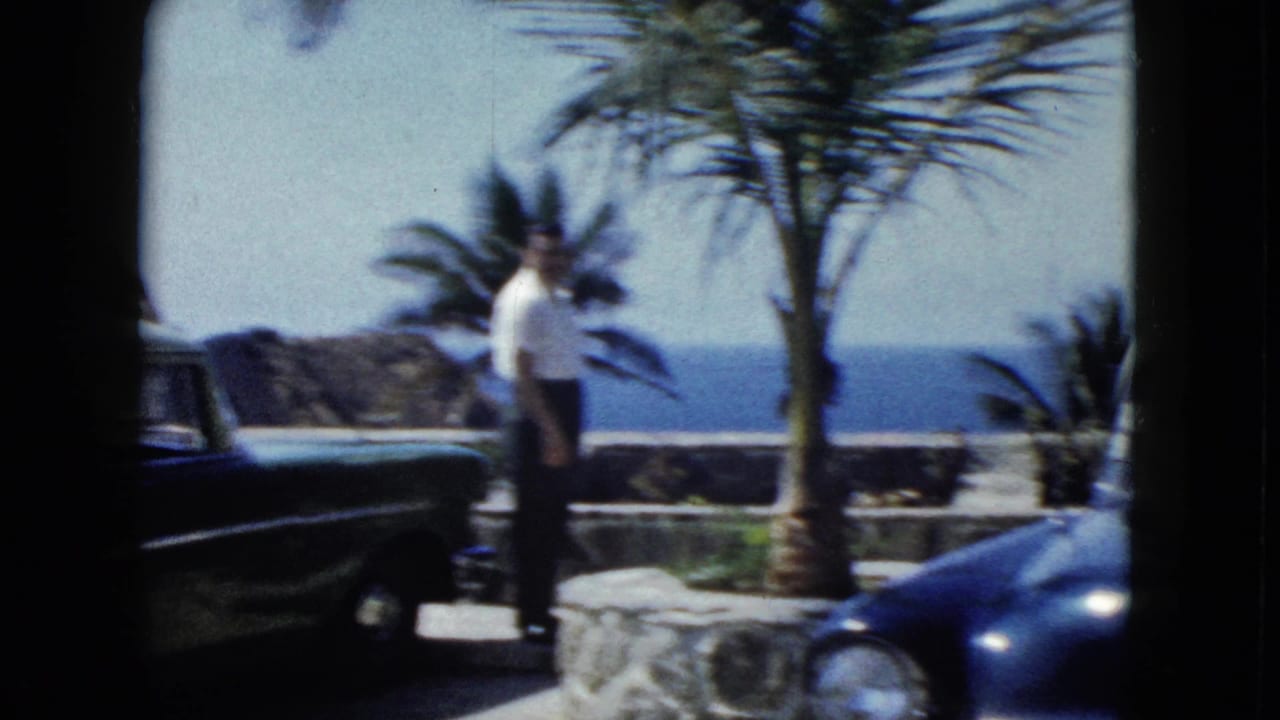 Same tourist, slightly repositioned, on a coastal stone terrace in Acapulco circa 1962; dark blue car prominent in foreground, Pacific Ocean and rocky cliffs behind.