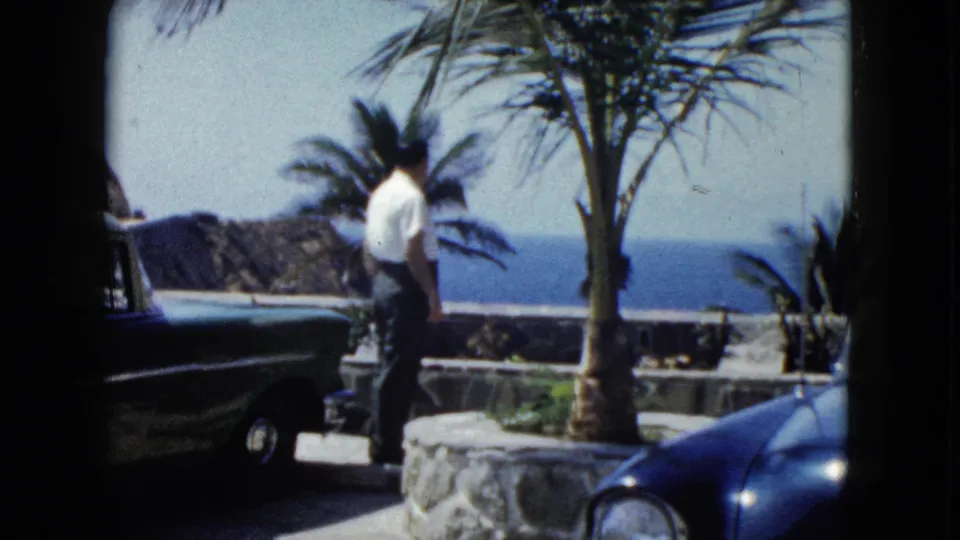 Tourist turns away from camera on a stone coastal terrace in Acapulco circa 1962, dark blue early-1960s car in foreground, palm tree and Pacific bay behind.