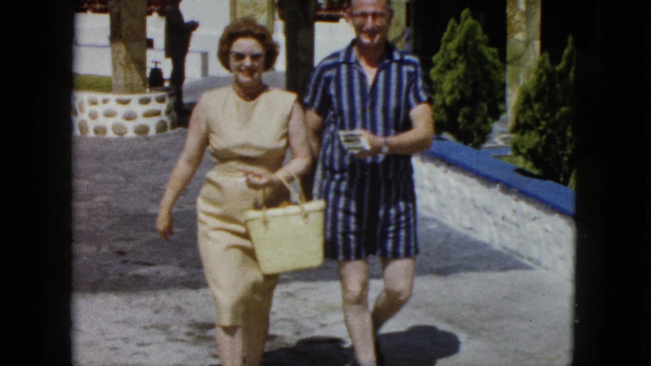 A woman in a 1960s sleeveless sheath dress with sunglasses and a man in striped resort wear walk together along a stone path at what appears to be an Acapulco resort, circa 1962.