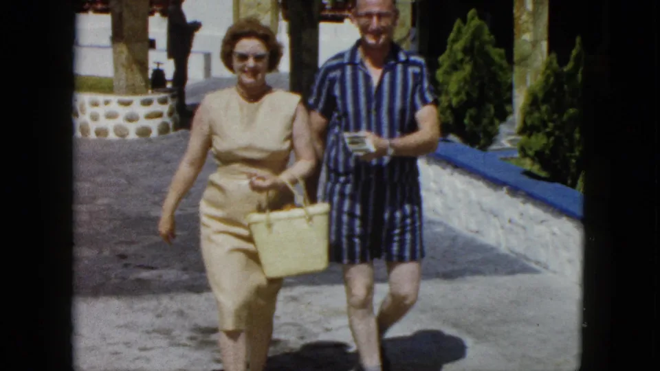 A woman in a 1960s sleeveless sheath dress with sunglasses and a man in striped resort wear walk together along a stone path at what appears to be an Acapulco resort, circa 1962.