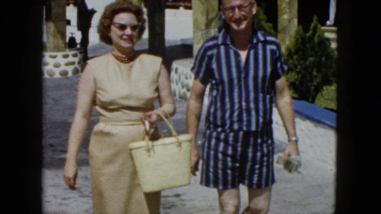 A woman in a beige sheath dress with pearl necklace and cat-eye sunglasses and a man in striped resort wear smile while walking along an Acapulco resort colonnade, circa 1962.