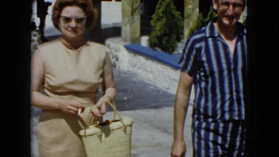 Close view of a woman in a sleeveless dress with sunglasses and pearl necklace and a man in striped resort wear, both smiling at the camera at what appears to be an Acapulco resort, circa 1962.