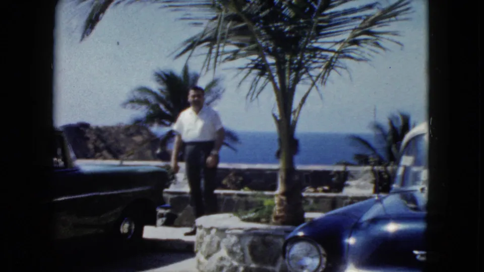 A man in a white shirt stands beside what appears to be a late-1950s or early-1960s dark-colored automobile, with a palm tree and coastal ocean view behind him, likely Acapulco, circa 1962.