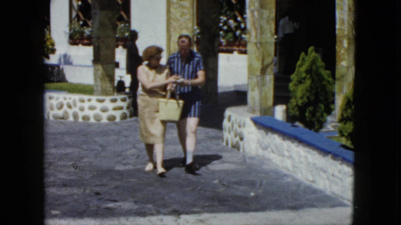 Middle-aged tourist couple strolling a resort courtyard in Acapulco, Mexico, circa 1962, on 8mm film; woman in beige sheath dress, man in blue striped shirt, yellow wicker basket in hand.