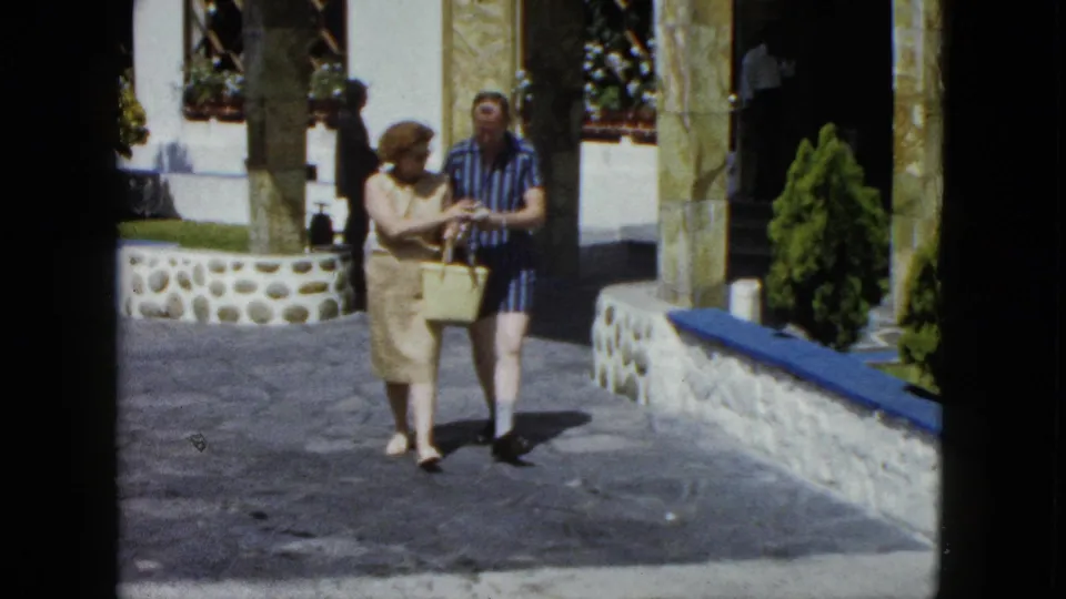 Middle-aged tourist couple strolling a resort courtyard in Acapulco, Mexico, circa 1962, on 8mm film; woman in beige sheath dress, man in blue striped shirt, yellow wicker basket in hand.