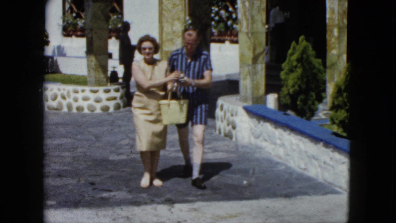 Tourist couple at a Mexican resort, early 1960s; woman in sunglasses and beige dress holds a wicker basket as both appear to examine its contents, stone colonnade with blue trim behind.