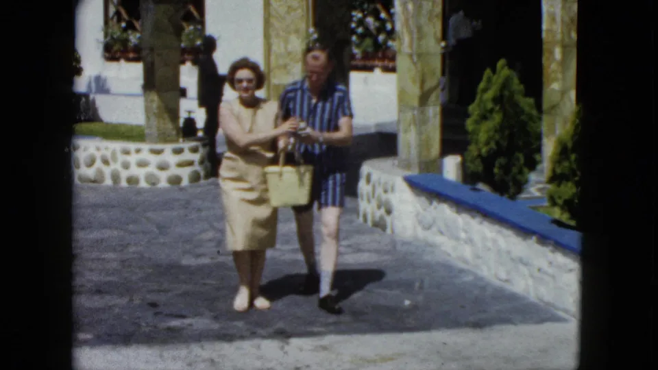 Tourist couple at a Mexican resort, early 1960s; woman in sunglasses and beige dress holds a wicker basket as both appear to examine its contents, stone colonnade with blue trim behind.