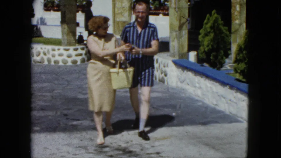 Smiling couple in 1960s resort attire crossing a sunlit Mexican resort courtyard; man in striped shirt grins as woman looks down at yellow wicker basket, captured on 8mm film.