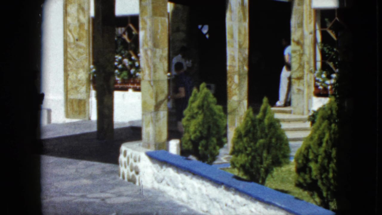 Wide courtyard of an ornate building with stone columns, blue-painted stone ledge, conical topiary shrubs, and background figures.